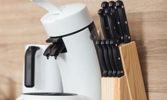 A modern kitchen corner with a white coffee machine and a wooden block with knives. The countertop is black and the background is made of light wood.