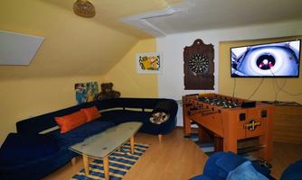 A cozy room with a blue sofa, a table, and a foosball game. A TV is hung on the wall and a dartboard is also visible.