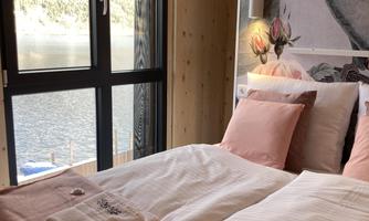 A cozy bedroom with two beds and soft, pink towels. The large window offers a view of a lake and the mountains in the background.