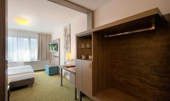 A modern hotel room with two beds, a large window, and a television. The decor is minimalist and friendly, with wooden and color accents.