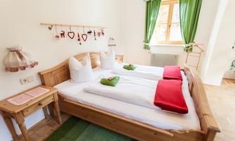 A cozy bedroom with two beds decorated with white bedspreads and red towels. The windows are adorned with green curtains, and the room has a warm, inviting character.