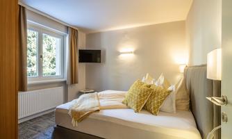 A cozy bedroom with a large bed and decorative pillows. A window provides plenty of natural light, and there is a television on the wall.