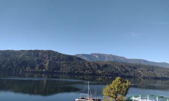 A calm lake surrounded by green hills and mountains. In the foreground, boats and a small marina can be seen.