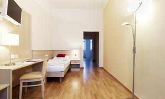 A simple, modern room with a single bed, a desk, and a television. The floor is wooden and there is a floor lamp in the room.