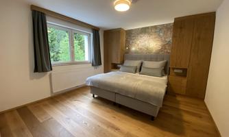 A modern bedroom with a large bed and wooden floor. The walls are plain and the windows let in plenty of daylight.