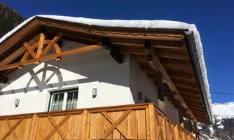 A cozy chalet with a wooden balcony and snow-covered roof. The sky is clear and blue, surrounded by mountains.