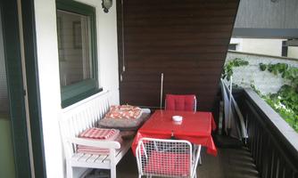 A cozy balcony with a red table and white chairs. The wall is made of wood and there are plants in the background.
