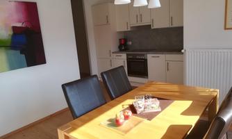 A bright dining area with a wooden table and black chairs. In the background, there is a modern kitchen with white cabinets.
