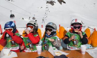 Four children in ski gear are sitting at a table and enjoying snacks in the snow. In the background, there are ski lifts and snow-covered mountains.