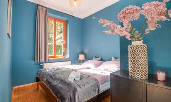 A cozy bedroom with blue walls and a large window. Soft blankets are on the bed and a bouquet of flowers is on the nightstand.