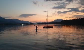 A man stands in the water with his arms raised in the air. The view shows a beautiful sunset over a lake, surrounded by mountains.