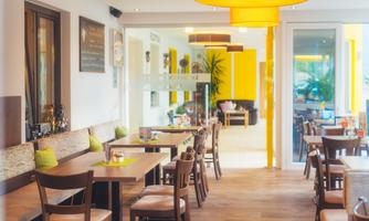 A modern restaurant with wooden tables and chairs. The walls are painted light and there are large windows that let in a lot of light.