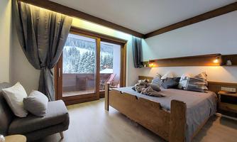 A cozy room with a large bed and a balcony overlooking snow-capped mountains. The wood paneling and soft lighting create a warm atmosphere.