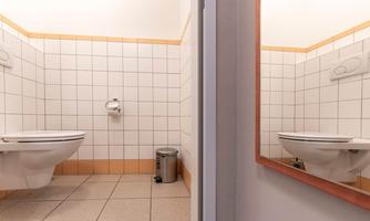 A simple bathroom with white tiles and a sink. A toilet and a garbage can are visible.