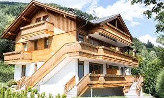A modern wooden house with several balconies in a green environment. The sky is clear and the mountains are visible in the background.