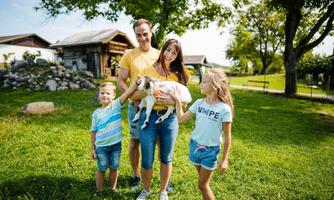 A family stands in the greenery holding a small animal. In the background, a farmhouse and trees are visible.