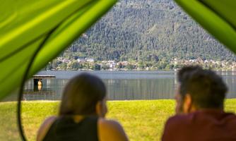 A couple is sitting in the tent, looking at a calm lake with mountains in the background. The scene radiates tranquility and a connection to nature.