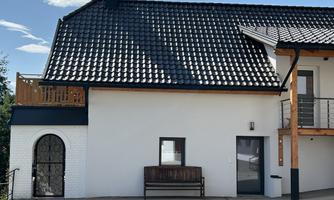 A modern house with a dark shingle roof. The façade is painted light and there is a wooden terrace with seating.