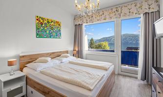 A modern bedroom with a large bed and a colorful painting. Large windows offer a view of the mountains and the sky.