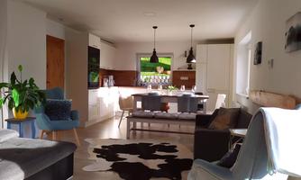 A modern, cozy living room with bright colors and an open kitchen. The space is friendly and inviting, with several seating options and a large dining table.