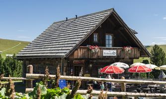 A cozy wooden house with a shingled roof. In front of the house are sunny terraces with red umbrellas.