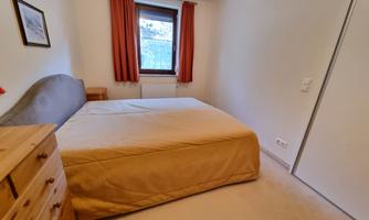 A simple bedroom with a large bed and a dresser. The walls are light, and there are orange curtains in front of the window.