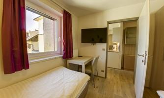 A small, bright room with a single bed and a desk. The room has a window with curtains and a television on the wall.