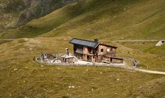 A rustic wooden house on a green meadow surrounded by mountains. The sky is clear and sunny.