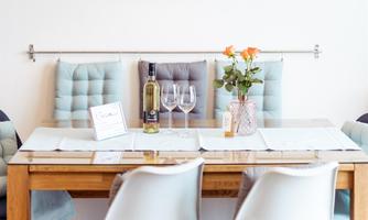 A modern dining area with a wooden table and stylish chairs. On the table are wine and a flower vase, while a lamp illuminates the room.