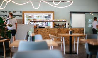 A modern bar in a bright room with wooden details. In the background, two people are ordering drinks.