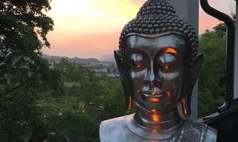 A Buddha statue made of silver stands on a windowsill, illuminated by a small candle. In the background, a beautiful sunset is visible with trees and a gentle landscape.