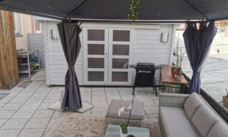 A cozy outdoor area with a sofa and a table under a tent roof. In the background, there is a grill and a wooden structure.