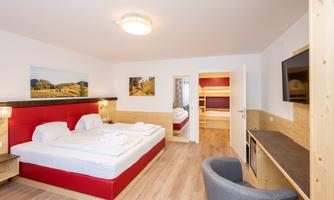 A modern hotel room with a double bed and a small table. In the background, another room with bunk beds is visible.