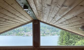 A room with a wooden ceiling and large windows offers a view of a tranquil lake and surrounding green hills. The atmosphere is inviting and cozy.
