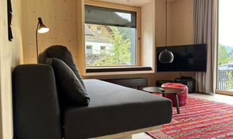 A modern living room with a cozy sofa, a television, and a colorful rug. Large windows offer a view of nature.