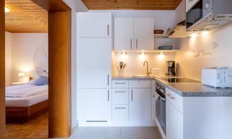 A modern kitchen with white cabinets and a granite countertop. In the background, a cozy bedroom with a double bed can be seen.