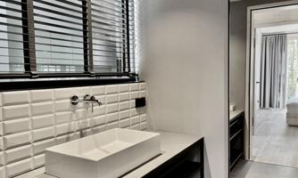 A modern bathroom with a stylish sink and a large mirror. Daylight streams through the window, and in the background, you can see a living area with curtains.