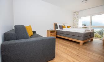 A modern bedroom with a double bed and a cozy sofa. Large windows provide plenty of natural light and a view outside.