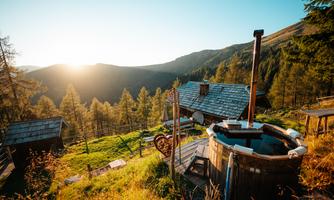 A picturesque mountain landscape with a wooden house and a warm spring in the foreground. The sun rises behind the mountains, bathing the scene in warm light.