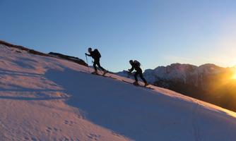 Two skiers ascend a snow-covered piste at sunset. Mountains can be seen in the background.