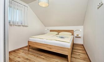 A cozy bedroom with a double bed and light wooden floor. The room has a simple decor and a window with curtains.