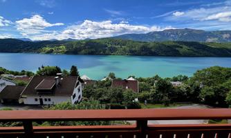 A beautiful view of a clear lake, surrounded by green hills and a blue sky. In the foreground, some houses and trees are visible.