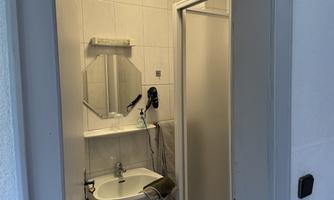A small bathroom with a shower, sink, and mirror. The walls are tiled and there is bright lighting.