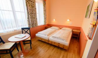A cozy room with two single beds and wooden flooring. There is a small table and chairs as well as large windows with curtains.