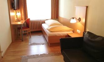 A simple bedroom with a single bed, a desk, and a television. The room has large windows that provide natural light.