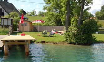 A beautiful garden by the water with a wooden terrace. Two people are relaxing under a sunshade.