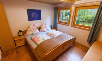 A cozy bedroom with a large double bed and colorful pillows. Large windows offer a view of the garden and provide plenty of daylight.