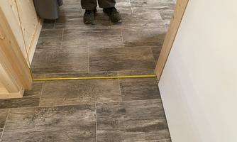 A floor with a wood look in a room. A person is standing in the doorway while a measuring tape is laid out on the floor.