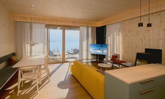 A modern living room with large windows and a view of the water. There is a comfortable yellow sofa, a dining area, and a television.