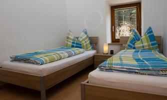 A bright room with two single beds and colorful bedcovers. There is a window and a bedside table with a lamp.